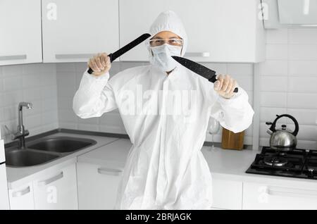 Homme en costume de protection blanc et en masque médical tient un couteau deux et fixe dans un appareil photo dans la cuisine blanche à la maison pendant le coronavirus. Séjour Banque D'Images