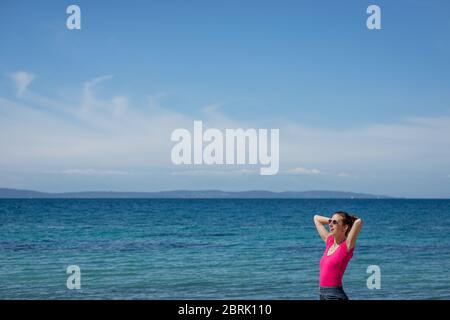 Jeune femme gaie en chemise rose penchée la tête sur ses mains derrière elle comme elle se tient au bord de la belle mer bleue. Banque D'Images