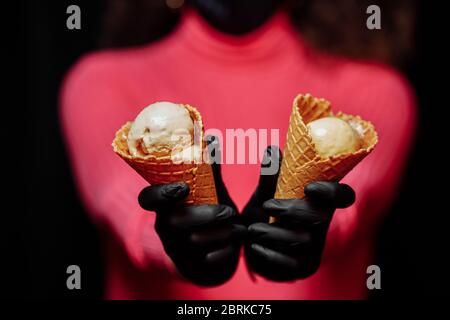 Les mains en gants de protection noirs maintiennent un cône à gaufre avec de la crème glacée. Protection contre le coronovirus. Concept de vente de crème glacée pendant la quarantaine Banque D'Images