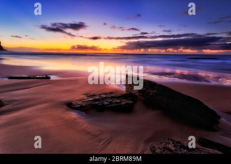 Rochers en grès foncé sur une plage de sable de Sydney sur la côte Pacifique au lever du soleil face à l'est. Banque D'Images