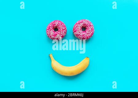 Deux beignets et une banane sous forme de visage souriant sur fond bleu, vue de dessus, plat Banque D'Images