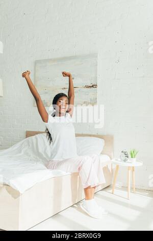 bonne fille afro-américaine en pyjama s'étirant avec les mains levées tout en étant assis sur le lit Banque D'Images