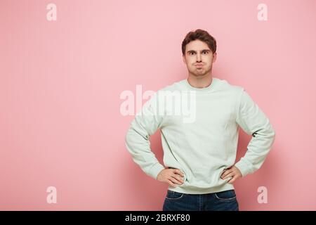 homme ennuyé qui puait les joues en se tenant debout avec les mains sur les hanches sur fond rose Banque D'Images