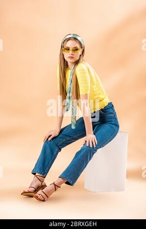 belle fille blonde en jean, t-shirt jaune, lunettes de soleil, sandales à talons et foulard en soie assis sur un cube blanc sur beige Banque D'Images