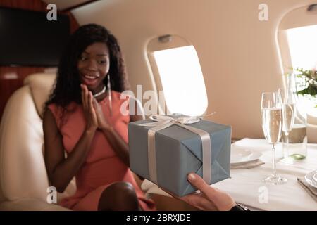 vue rognée de l'homme présentant une boîte cadeau à une femme afro-américaine surprise en avion privé Banque D'Images