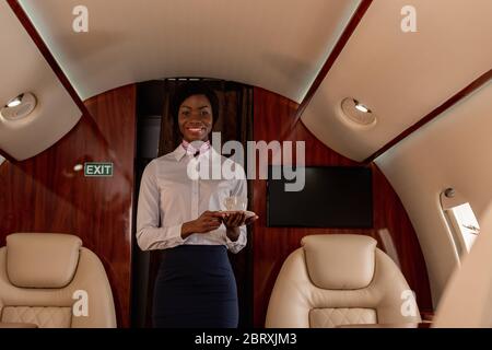 hôtesse afro-américaine souriante et attrayante regardant la caméra tout en tenant une tasse de café dans un jet privé Banque D'Images