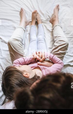 Une petite fille se repose sur la poitrine de sa mère dans le lit comme ils tiennent les mains ensemble. Maman l'enveloppe dans ses bras comme un signe d'amour Banque D'Images