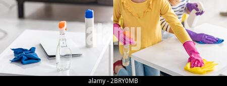 Collage de chiffon, bouteilles et ordinateur portable sur table basse et femme avec fille essuyant table basse dans le salon, photo panoramique Banque D'Images