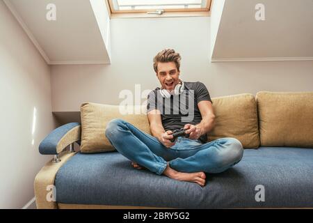 KIEV, UKRAINE - 13 AVRIL 2019: Homme excité dans des écouteurs sans fil sur le cou jouer au jeu vidéo tout en étant assis sur un canapé avec des jambes croisées Banque D'Images