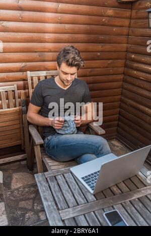 vue en grand angle du beau jeune homme triant tout en s'asseyant près d'un ordinateur portable sur le balcon Banque D'Images