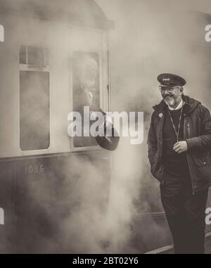 Vue monochrome d'un ouvrier de train à vapeur souriant sur une plate-forme qui discute avec l'inspecteur de billets par la fenêtre de quitter un train britannique d'époque. Un coup de vapeur ! Banque D'Images