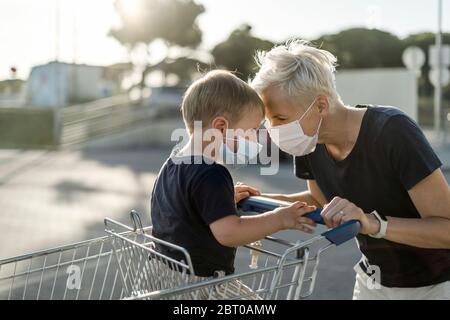 Mère jouant avec joie avec un enfant assis dans un chariot. Les deux portent un masque de protection avant d'entrer dans un supermarché. Banque D'Images