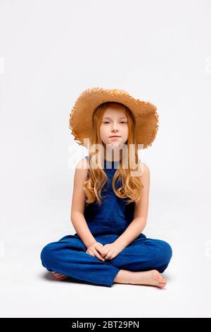 fille à cheveux rouges assise dans une combinaison bleue et un chapeau de paille, jambes croisées Banque D'Images