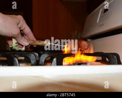 Une main tenant un folk et rôtisant deux hot dogs sur le brûleur d'un four à gaz. Banque D'Images