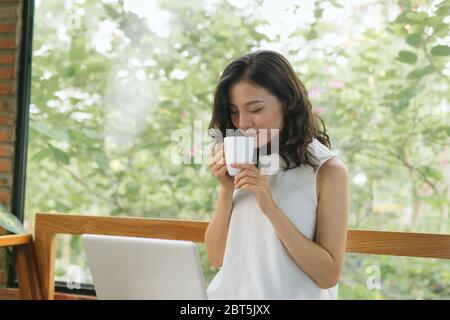 Femme indépendante tenant une tasse à café et utilisant un ordinateur portable sur une table dans un café-restaurant Banque D'Images