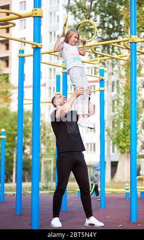 Un père heureux soutient sa fille pendant qu'elle est suspendue sur les barres de sport à l'extérieur. Entraînement en famille. Fond urbain Banque D'Images