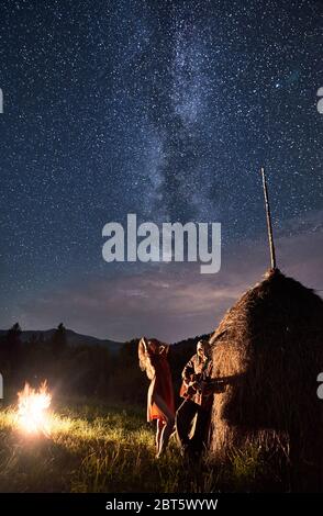 Couple romantique ayant un repos dans les montagnes, une femme dansant à côté du feu et l'homme jouant la guitare se pencha sur un rick de foin sec sous le ciel plein d'étoiles avec la voie lactée, longue exposition Banque D'Images