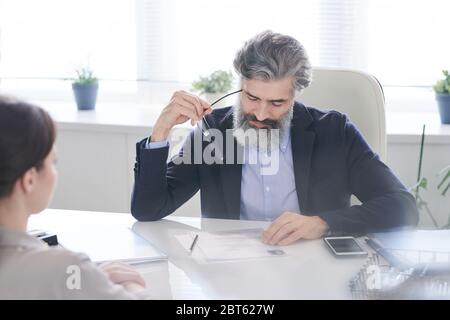 Manager d'une heure mâle pensive et âgée tenant des lunettes par le temple tout en lisant les points de CV de la candidate assise devant lui Banque D'Images
