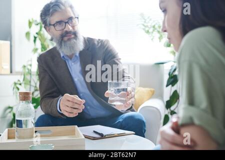 Conseiller senior avec barbe passant un verre d'eau à sa jeune femme en difficulté pendant la conversation par table au bureau Banque D'Images