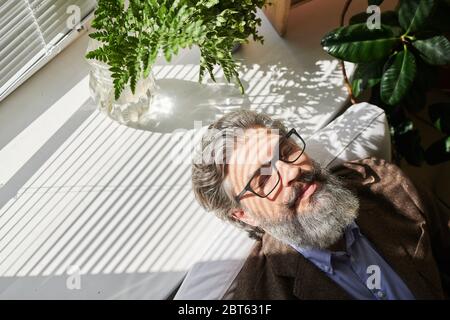 Heureux homme d'affaires mature et détendu ou autre professionnel avec cheveux gris et barbe mettant sa tête sur le rebord de fenêtre après le travail au bureau Banque D'Images