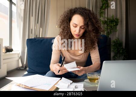 Jeune femme d'affaires sérieuse avec de longs cheveux ondulés regardant par les factures de paiement tout en étant assis sur un canapé devant un ordinateur portable Banque D'Images