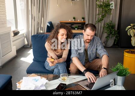 Jeune femme avec carte en plastique assise sur un canapé à côté de son mari devant un ordinateur portable tout en regardant les deux exposés pendant les achats en ligne Banque D'Images