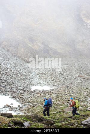 Vue latérale des randonneurs amis avec des sacs à dos marchant sur une colline herbeuse avec une énorme montagne brumeuse sur fond. Deux hommes avec bâtons de randonnée randonnée longue distance. Concept de voyage, tourisme, randonnée Banque D'Images