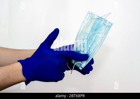 main dans un gant médical bleu tient un masque d'hygiène bleu une autre main avec le pouce vers le haut sur un fond blanc Banque D'Images