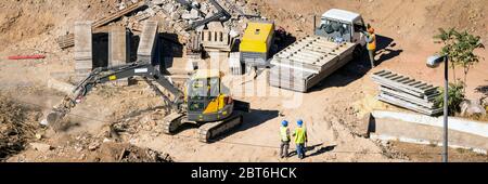 Un ouvrier du chantier portant une combinaison bleue et une veste de sécurité jaune haute visibilité debout et observant la tranchée de creusage de godet sur une roquette sablonneuse Banque D'Images