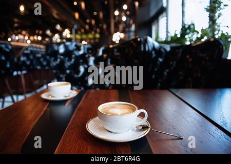 deux tasses de café sont sur la table, restaurant, meubles bruns, lumières tamisées, une tasse blanche et une cuillère en métal sur une assiette, gros plan, cappuccino dans une tasse, Banque D'Images