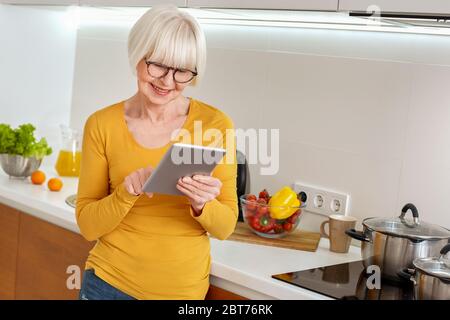 Retraite. Femme senior dans des verres debout à la cuisine de navigation Internet à la recherche de recette sur tablette numérique sourire heureux Banque D'Images