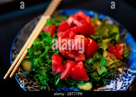 Macro-gros plan de plat de salade de tomates vertes avec des baguettes et des légumes coupés hachés sur une plaque en céramique bleue traditionnelle dans le ryok du restaurant gastronomique japonais Banque D'Images