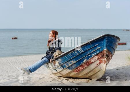 Bonne jeune femme qui profite du soleil de printemps, assise sur un vieux canot en bois sur une plage de sable balançant ses jambes et riant