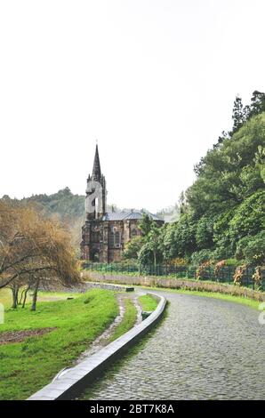 Chemin pavé au bord du lac entouré d'arbres verts. Chapelle de Nossa Senhora das Vitorias à Furnas, Açores, Portugal en arrière-plan. Couvert, jour de pluie, ciel blanc. Photo verticale. Banque D'Images