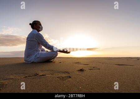 méditant homme dans le masque médical dans lotus poser sur la plage, homme portant le masque chirurgical et les vêtements de yoga blancs avec la coiffure de haut nœud au lever du soleil Banque D'Images