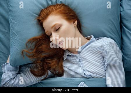 Belle fille à cheveux rouges en pyjama rayé dort dans un lit bleu. Cadre sur le dessus. Banque D'Images