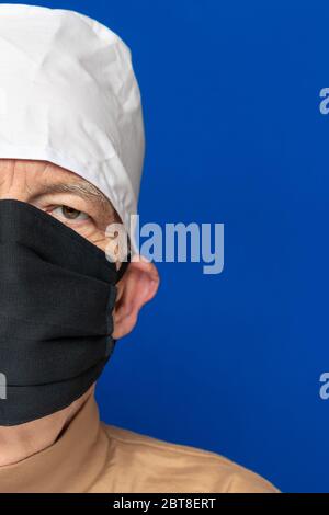 Demi-visage adulte senior médecin habillé noir masque chirurgical, blanc capuchon médical, debout sur fond bleu et regardant pierly la caméra Banque D'Images