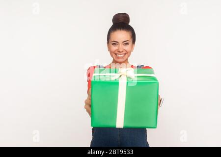 Portrait de bonne joyeuse jolie fille avec un pain de cheveux tenant un énorme cadeau emballé, donnant le cadeau à l'appareil photo et souriant, félicitant pour les vacances, offeri Banque D'Images