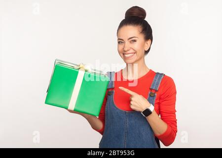 Regardez le cadeau ! Portrait de bonne fille joyeuse positive avec un petit pain dans des salopettes en denim pointant vers la boîte à présent enveloppée et souriant, félicitant sur h Banque D'Images