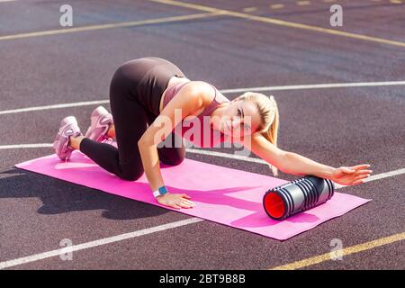 Athlète sportive blonde femme entraînement sur tapis en extérieur jour d'été, utilisant le masseur à rouleau en mousse pour les douleurs de dos, étirant les muscles de main et de colonne vertébrale, faisant fasc Banque D'Images