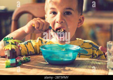 Le garçon 4 ans mange du porridge. Table enfant. Le concept de l'indépendance de l'enfant. Le garçon est déjeunant avec un appétit sur l'arrière cuisine Banque D'Images