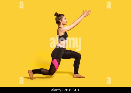 Vue latérale, petite femme mince avec cheveux petits pains dans des vêtements de sport serré faire l'exercice de fente sportive, debout un genou et levant les mains, réchauffant l'entraînement mus Banque D'Images