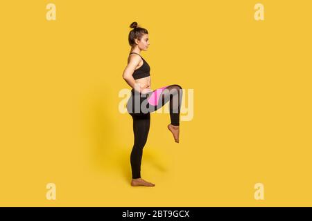 Vue latérale, femme athlétique mince avec cheveux petits pains dans les vêtements de sport serré faisant du sport debout avec un ascenseur de jambe, entraînement de cuisses, échauffement des muscles d'entraînement. Banque D'Images