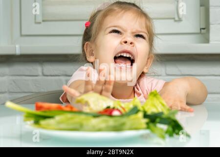 une fille de 4 ans repousse émotionnellement une assiette de légumes d'elle-même. manque de fruits et de légumes dans l'alimentation des enfants Banque D'Images