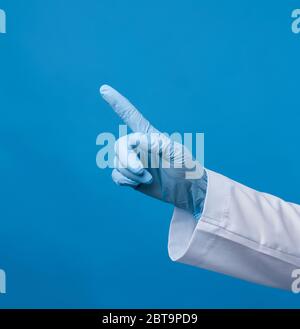 femme medic en manteau blanc, portant des gants stériles bleus, montrant un geste de la main indiquant le sujet, fond bleu Banque D'Images