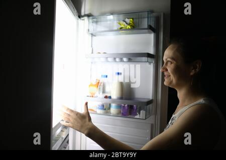 Une femme souriante la nuit à la maison a ouvert le réfrigérateur Banque D'Images