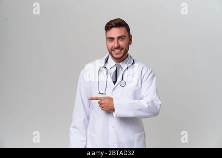 Beau moderne docteur blanc robe médicale dans studio gris de la publicité de fond de votre produit regardant l'appareil photo et pointant le doigt vers le positiv Banque D'Images