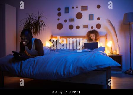 Portrait complet de jeunes couples mixtes utilisant des gadgets d'ordinateur dans le lit la nuit, espace de copie Banque D'Images