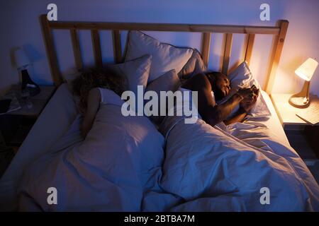 Au-dessus de la vue portrait de l'homme afro-américain utilisant un smartphone dans le lit la nuit tout en étant allongé à côté de la femme ou de la petite amie, espace de copie Banque D'Images
