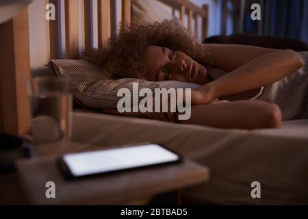 Portrait d'une jeune femme aux cheveux bouclés dormant calmement dans un lit la nuit avec un smartphone éclairé sur une table de nuit, espace de copie Banque D'Images